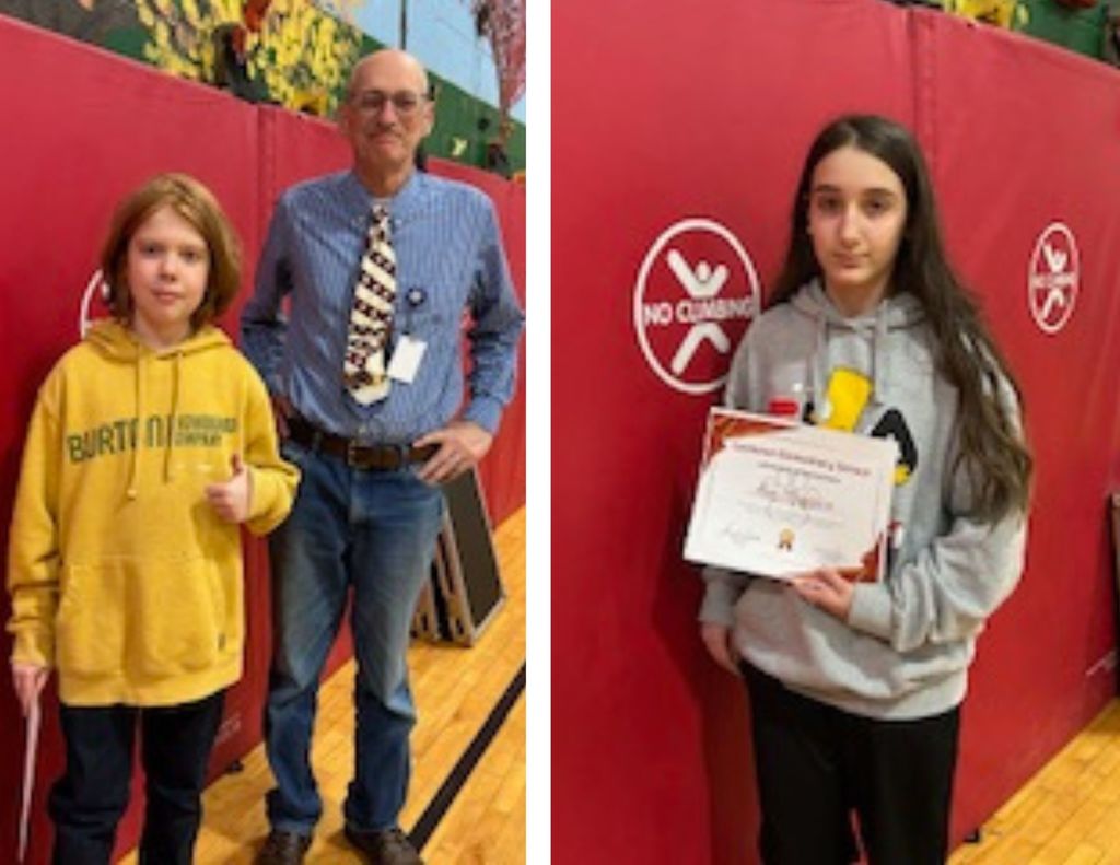 A student holds a certificate while standing next to a staff member in a school gym, posed in front of red wall mats.
