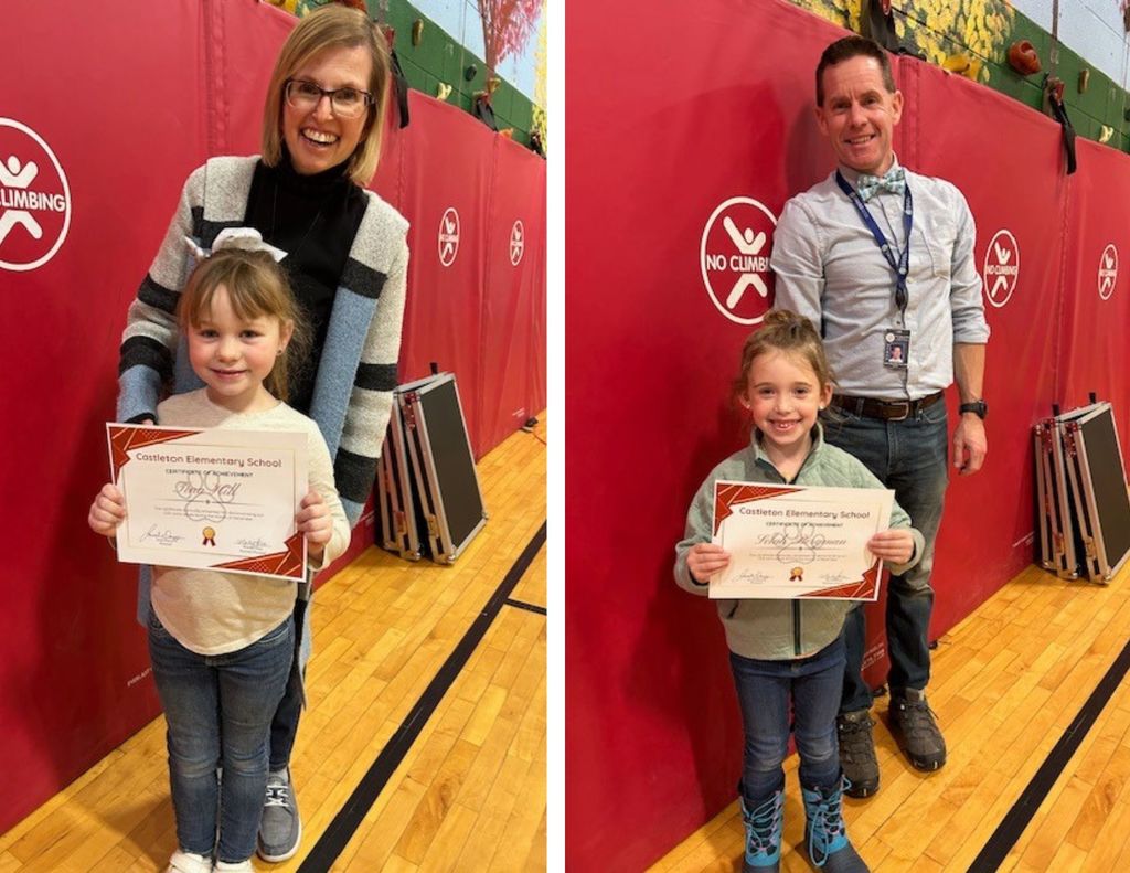 Two elementary students stand in a school gym holding certificates, each posing with a staff member against red safety mats.