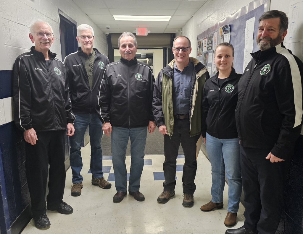 Seven Vermont Soccer Officials Association representatives stand together in a school hallway, wearing black VSOA jackets and smiling for a group photo.