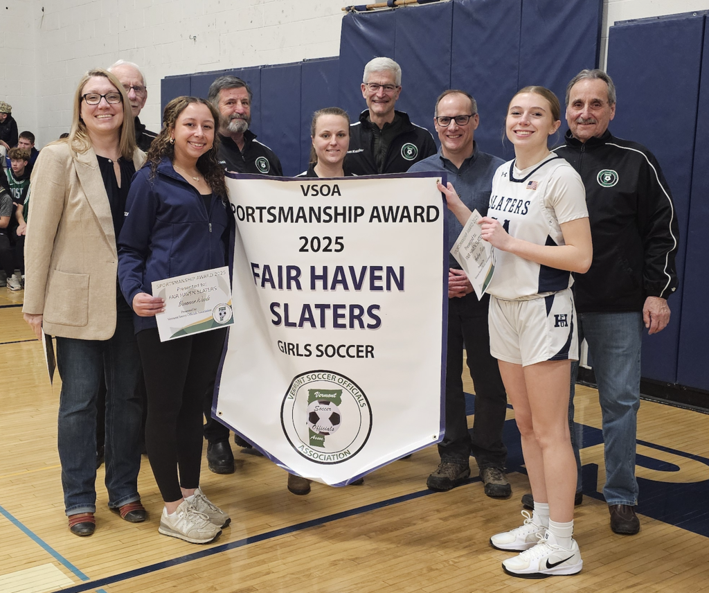 Fair Haven Slaters girls’ soccer player and coaches stand with Vermont Soccer Officials Association representatives holding a banner that reads “Sportsmanship Award 2025 – Fair Haven Slaters” during an indoor recognition ceremony.