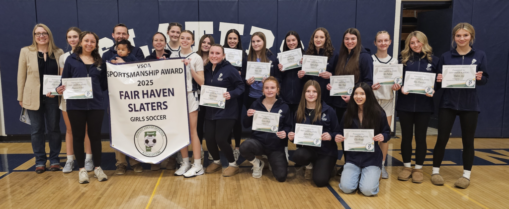 The Fair Haven Slaters girls’ soccer team, coaches, and staff pose in a gymnasium holding individual certificates and a large banner reading “VSOA Sportsmanship Award 2025 – Fair Haven Slaters Girls Soccer.”