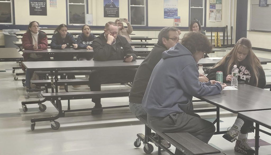 Community members and students sit at cafeteria tables during an evening school event, listening and reviewing materials.