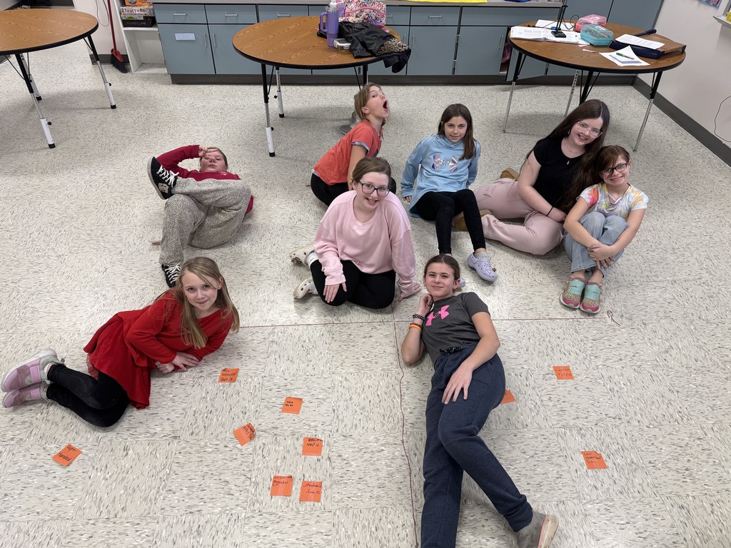 The same group of nine students from a different angle, sitting around the floor graph in a classroom. In this version, the students are more spread out and many are making silly faces or poses for the camera. The floor graph with its tape axes and orange sticky note data points remains the central focus of the room.