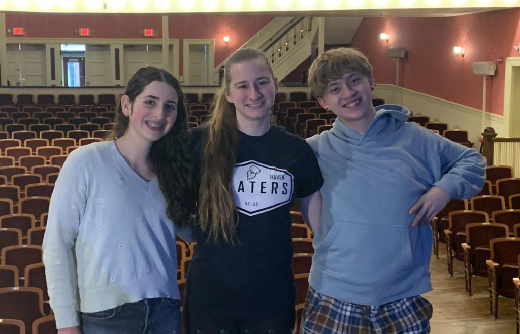 Three students stand smiling on a theater stage with rows of empty auditorium seats behind them, representing their school at the Vermont Drama Council Winter Meeting.