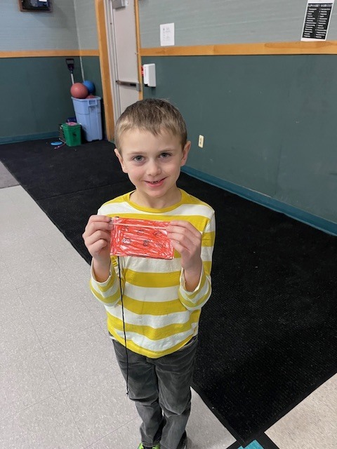 A young boy with light brown hair smiling at the camera, wearing a yellow and white striped shirt, holds up a small, rectangular red craft object with black string. He is standing on a black mat in a gymnasium or multi-purpose room with a grey floor and green and wood-paneled walls in the background.