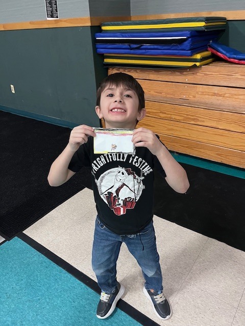 A young boy with a wide smile, wearing a black t-shirt with a "Frightfully Festive" graphic and blue jeans, holds up a small, rectangular handmade paper craft. He is standing in a gymnasium with wooden bleachers and a stack of colorful gym mats behind him.