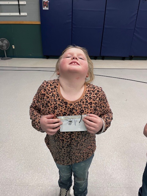 A young girl in a leopard-print shirt, with her head tilted back and smiling, holds a small white envelope-like paper craft with a string attached in front of her. She is standing on a speckled floor in a gymnasium, with blue padded mats and a basketball court line visible behind her.