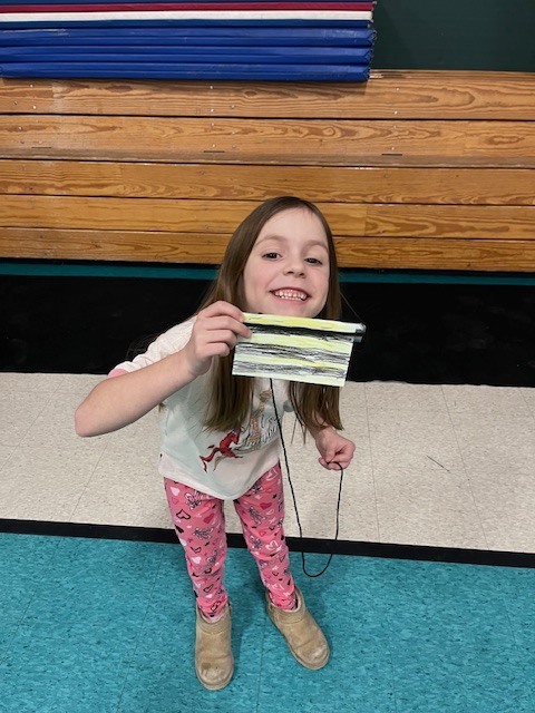 A young girl with long brown hair, wearing a white graphic t-shirt and pink heart-patterned leggings, leans forward and smiles at the camera. She is holding a small, handmade paper craft with yellow and black stripes, designed to look like a bee, which is attached to a black string. She stands in front of wooden bleachers and a stack of blue gym mats in a gymnasium.