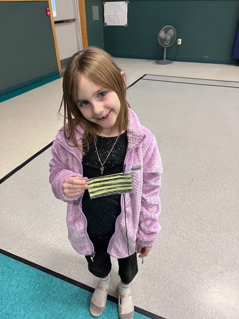 A young girl with long light brown hair, wearing a purple fuzzy zip-up jacket over a black glittery top and black pants, smiles at the camera. She is holding a small, rectangular handmade craft with yellow and black horizontal stripes. She stands in a school gymnasium on a light grey tiled floor with black and teal lines, with a doorway and a floor fan visible in the background.