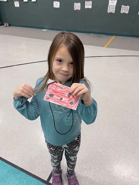 A young girl with long brown hair wearing a teal shirt and floral leggings holds a red paper craft with black dots, designed to look like a ladybug. 