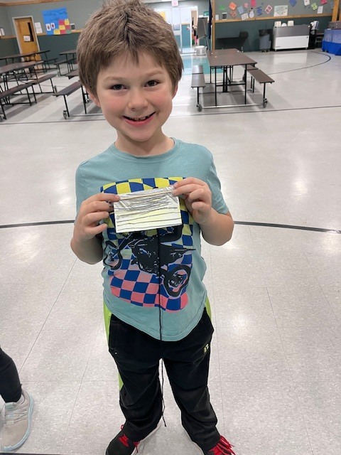  A young boy with messy brown hair and a bright smile wears a light blue t-shirt with a checkered graphic. He proudly holds up a handmade paper craft with yellow and black stripes. 