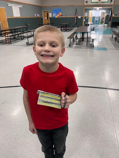 A young boy with a blonde bowl cut and an orange henley shirt stands in a gymnasium. He has a neutral expression while holding a paper craft decorated with yellow and black stripes. 