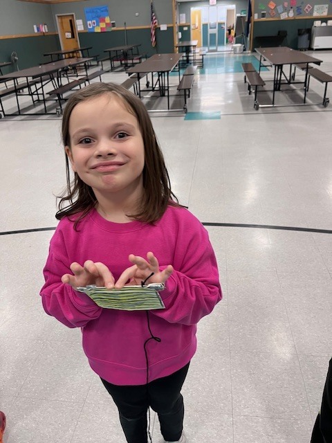 A girl in a bright pink sweatshirt stands in a school cafeteria, making a silly, tight-lipped face at the camera. She holds a yellow and black striped paper craft in both hands. 