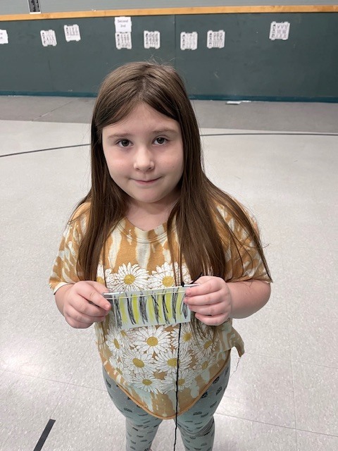 A young girl with long brown hair, wearing pink heart-patterned leggings and a white t-shirt, leans toward the camera with a big smile. She holds a handmade paper craft with yellow and black horizontal stripes. 