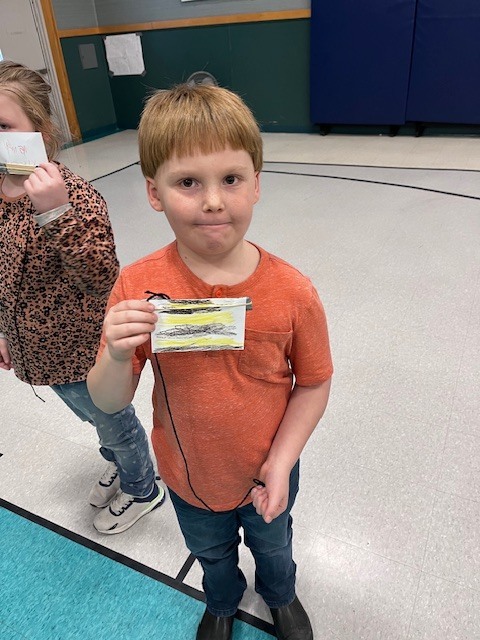 A young boy with a blonde bowl cut and an orange henley shirt stands in a gymnasium, holding a small, rectangular handmade paper craft with horizontal yellow and black stripes, resembling a bee, attached to a black string. He has a neutral expression. In the background, there are blue padded mats on a green wall, and a second child is partially visible on the left, holding a similar craft.