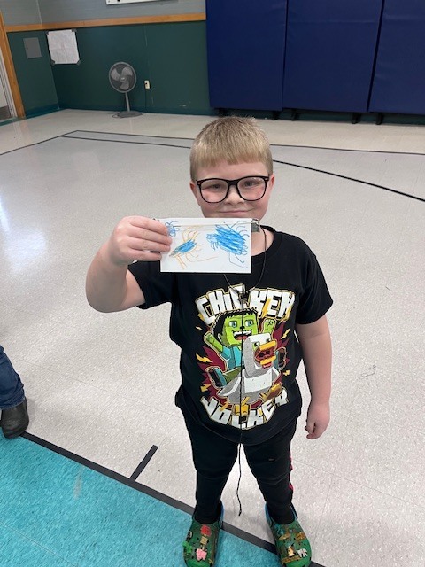 A young boy with a wide smile, wearing a black t-shirt with a "Frightfully Festive" graphic and blue jeans, holds up a small, rectangular handmade paper craft. He is standing in a gymnasium with wooden bleachers and a stack of colorful gym mats behind him.