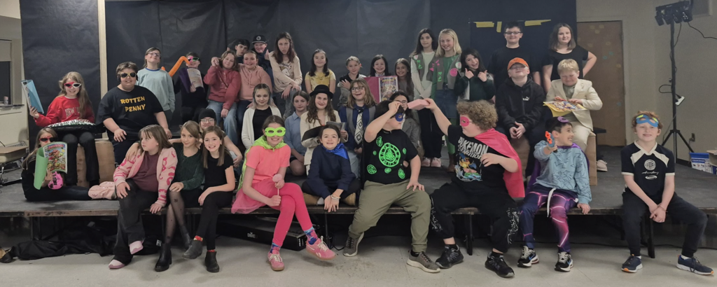 A large group of elementary students pose together on stage in colorful costumes, some wearing masks and holding comic books, smiling and showing playful expressions.