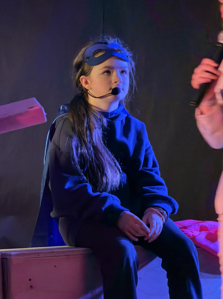 A seated student wearing a mask and headset microphone listens attentively while another person holds a microphone nearby, suggesting dialogue on stage.