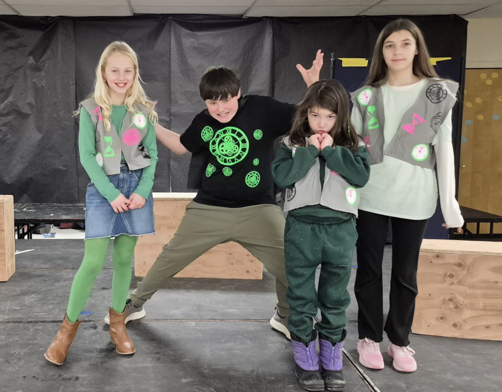 Four students stand in character wearing matching gray vests with bright patches, striking expressive poses as part of a dramatic scene.