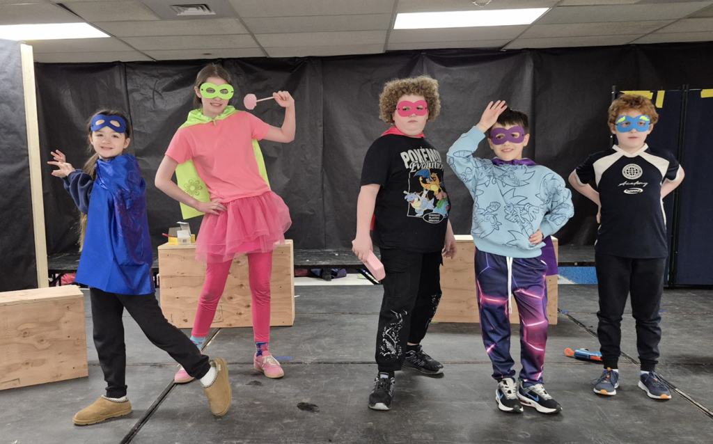 Five students pose as superheroes wearing colorful masks and capes, standing confidently on stage with hands on hips and playful stances.