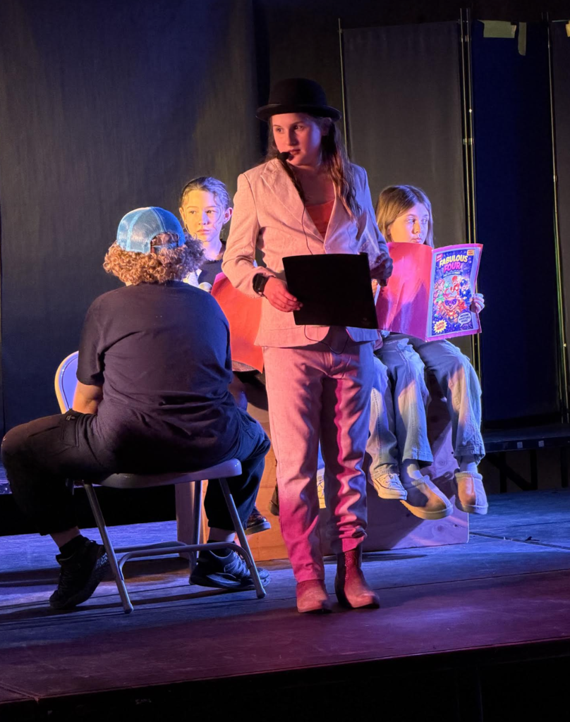 A student in a light-colored suit and hat holds a script while standing on stage, with other students seated behind holding comic books as part of the scene.