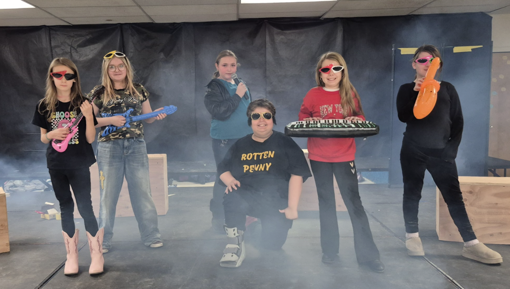 Six students stand on stage holding toy instruments and a microphone, dressed in casual costumes and sunglasses, posing like a rock band during the performance.
