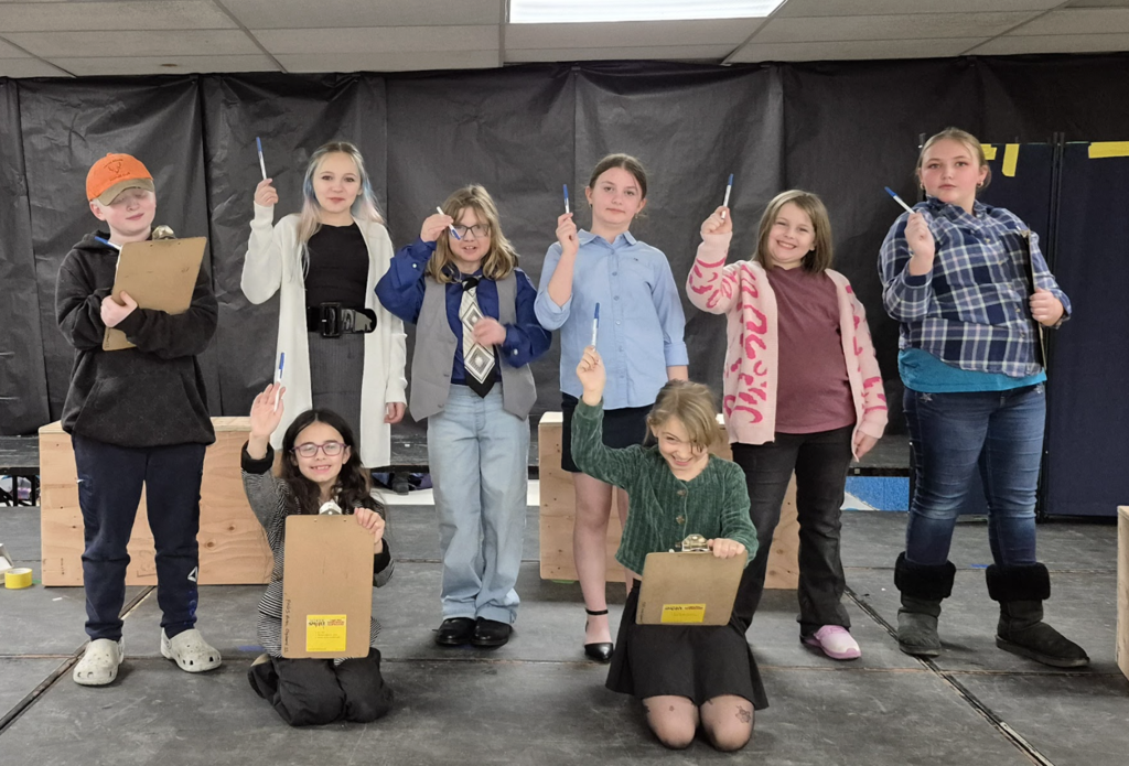A group of students stand and kneel on stage holding clipboards and pens, raising them in the air as if voting or making a decision in a scene.