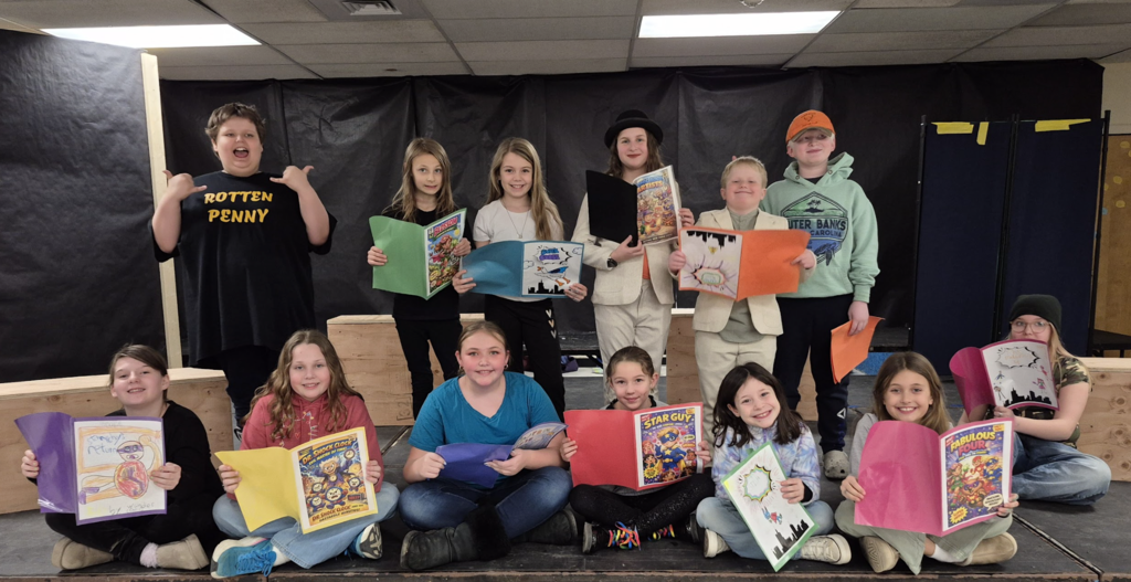 Students pose on stage holding handmade comic books and drawings, smiling proudly and displaying their artwork to the audience.
