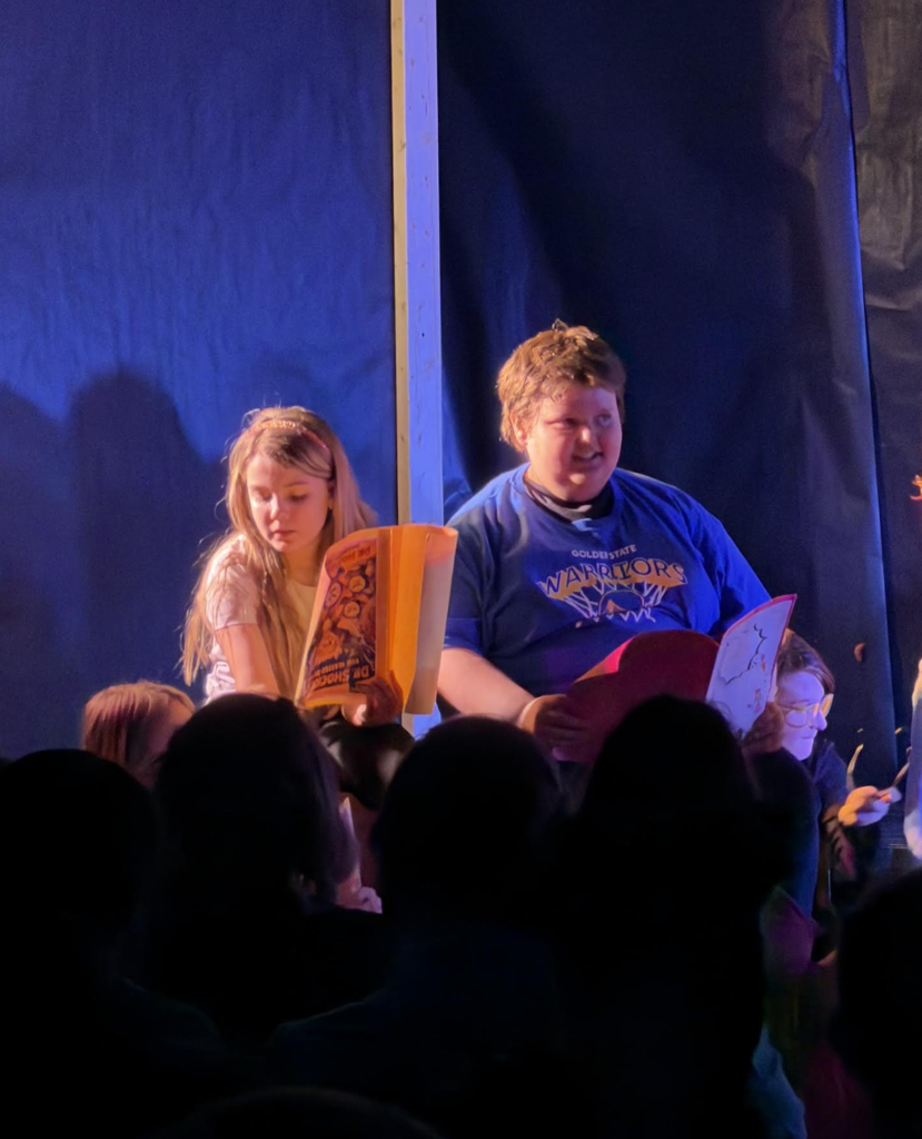 Two students sit on stage reading colorful comic books under stage lights, with the audience visible as silhouettes in the foreground.