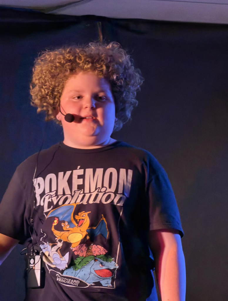 A student wearing a headset microphone and a Pokémon-themed shirt stands under stage lighting, speaking during a performance moment.