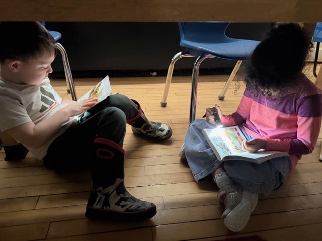 Two students sit on the floor under desks reading books with flashlights during a classroom activity.