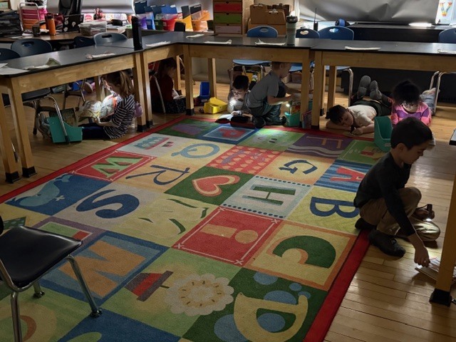 Wide view of classroom showing multiple students reading books by flashlight under tables and around a colorful rug.