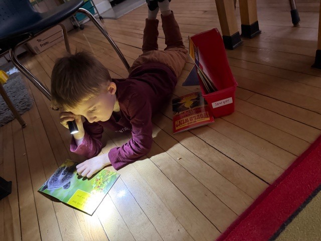 Young student lies on the classroom floor reading a picture book by flashlight, with a bin of books nearby.
