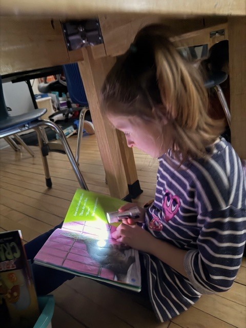 Student sits under a table reading a book by flashlight, illuminating the page on the floor.