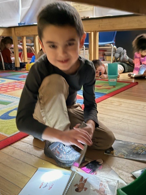 Student kneels on the floor under a table, smiling while reading a book illuminated by a flashlight.