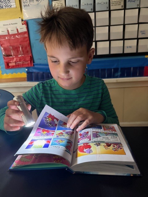 Student sits at a table reading a comic-style book by flashlight in a classroom.