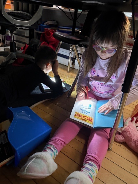 Student with glasses sits under a table reading a book by flashlight, while another student reads nearby.