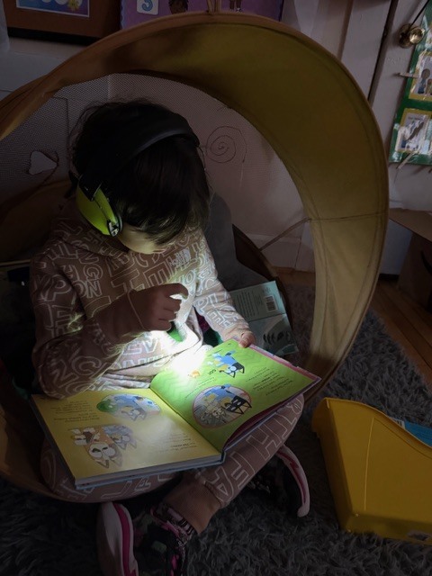 Student wearing headphones sits in a cozy reading chair, reading a picture book by flashlight.