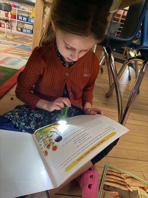 Student sits on the floor reading a book by flashlight, focused on the page beneath classroom desks.