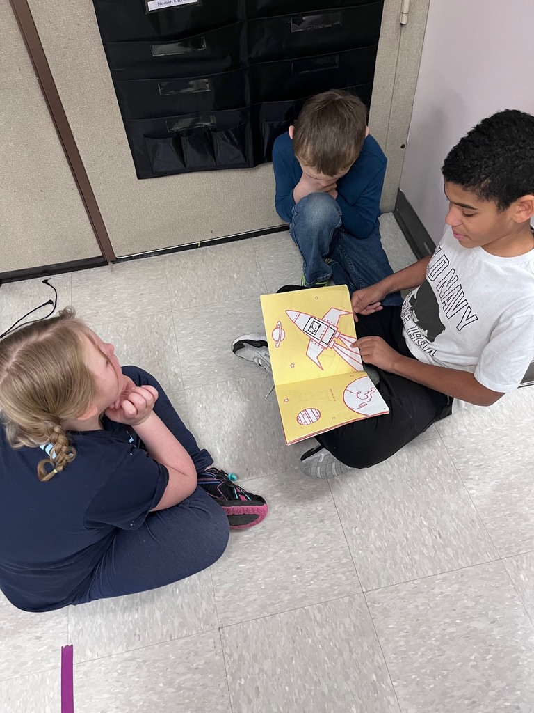  Three children are huddled together on a tile floor during a reading session. A boy in a white "Old Navy" t-shirt holds a yellow book featuring a simple illustration of a rocket ship and a planet, pointing to the page. A girl with blonde braids and a boy in a blue shirt watch closely from opposite sides.  i
