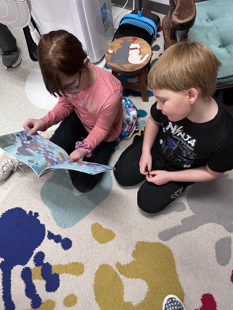  A girl in a pink long-sleeved shirt and a boy in a black "Ninja" t-shirt sit side-by-side on a handprint rug. The girl holds a thin picture book with blue-toned illustrations of a shark. In the background, there is a small wooden stool with cat-shaped cutouts on top. 