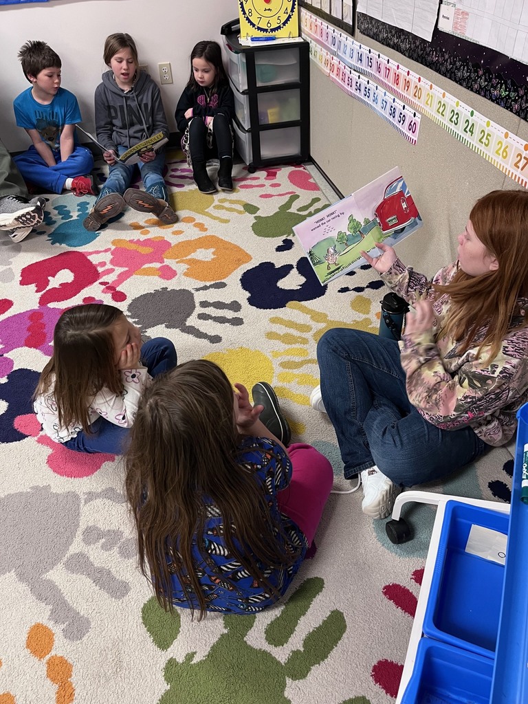 A high-angle shot of a classroom reading circle. A girl with long red hair sits on a colorful handprint rug, holding up a picture book featuring a red car to a group of five other children. The children are seated on the floor and a rug, listening intently. The background shows classroom decorations, including a number line and a yellow clock. 