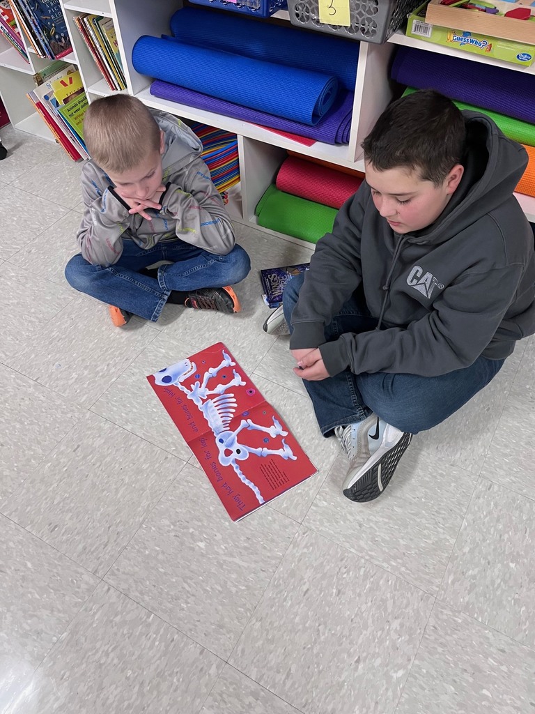  Two boys are seated cross-legged on a light-colored tile floor, looking down at an open picture book. The book shows a bright red page with a white skeleton of a dog. Behind them are classroom cubbies filled with colorful rolled-up yoga mats in blue, purple, green, and orange.  i