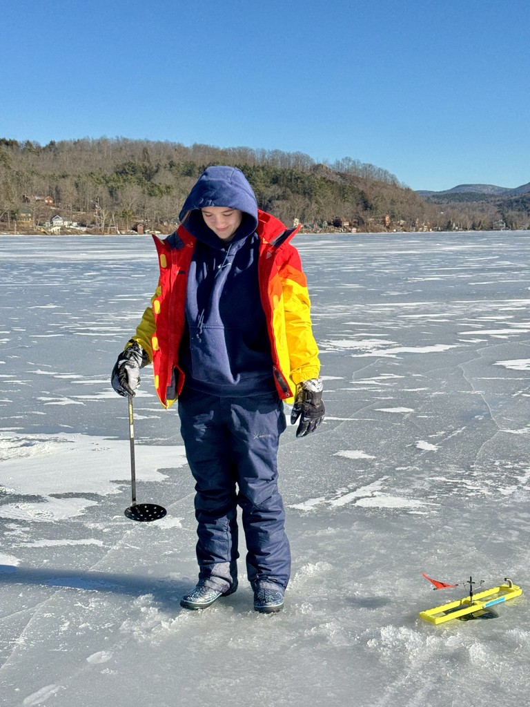 A student dressed in winter gear stands on a frozen lake holding an ice fishing skimmer, with ice fishing equipment on the ice nearby and hills in the background.