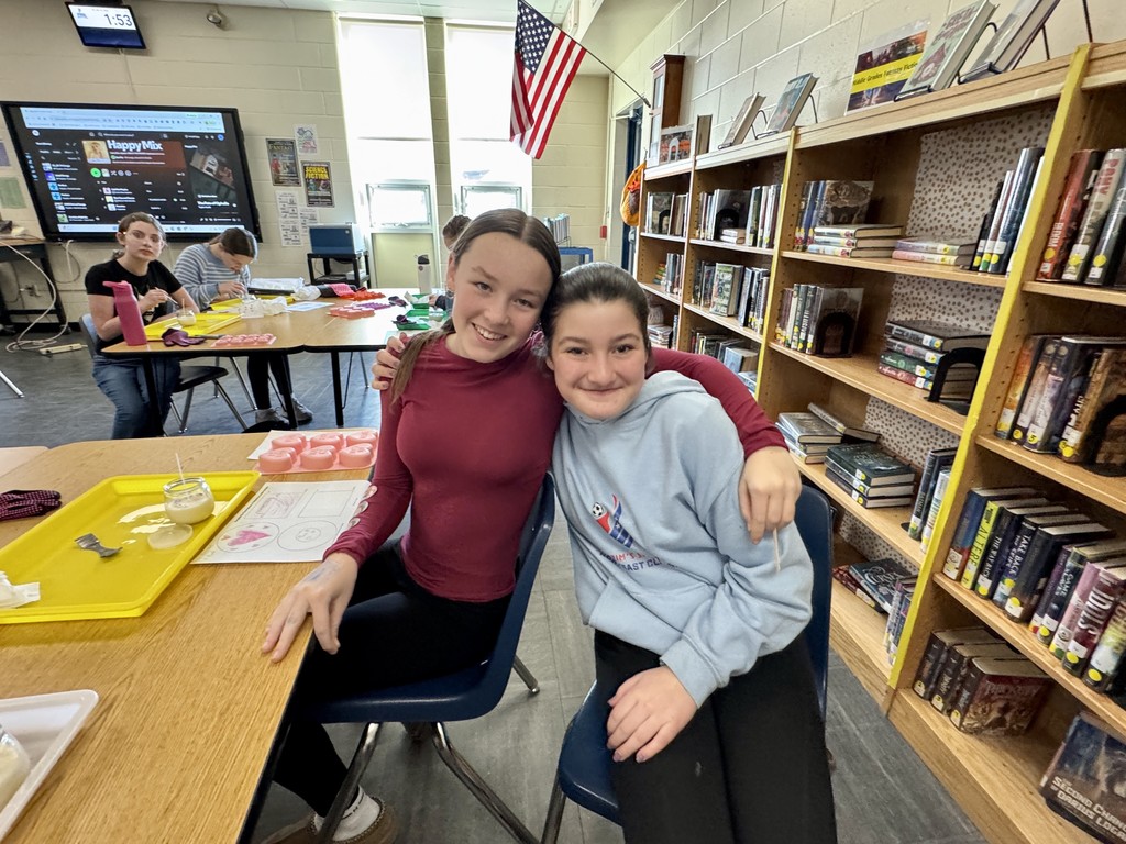 Two middle school students smile and pose with arms around each other at a table in the library while working on a hands-on activity, with other students crafting in the background.