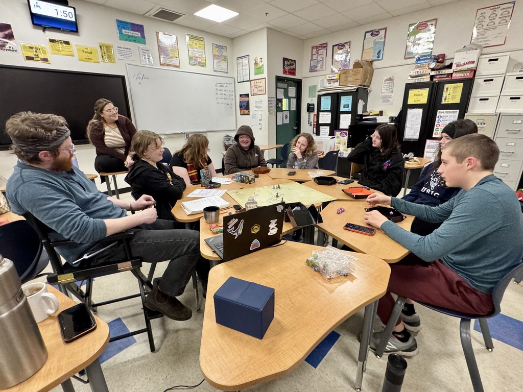 A group of middle school students sit around connected tables in a classroom playing a tabletop role-playing game, with game pieces, dice, and a large map spread out in the center.