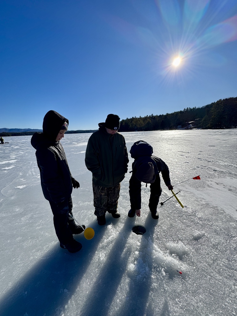 Three students bundled in winter clothing stand on a frozen lake looking down at an ice fishing hole, holding ice fishing tools under a bright sunny sky.