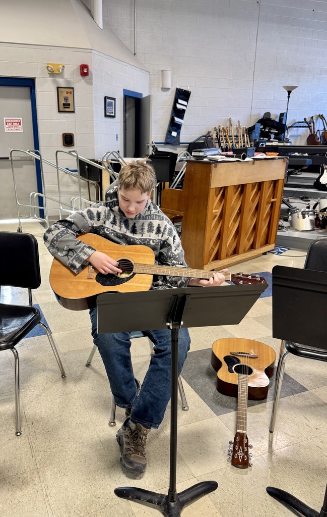 A student sits in a music room playing an acoustic guitar while reading from a music stand, with another guitar resting on the floor beside them.