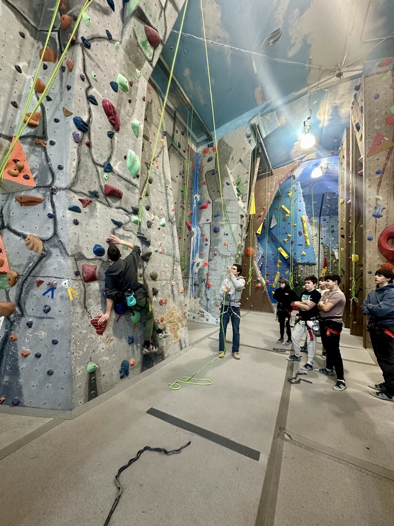 A student climbs an indoor rock wall while secured by a rope, as classmates and an instructor watch from below in a climbing gym.