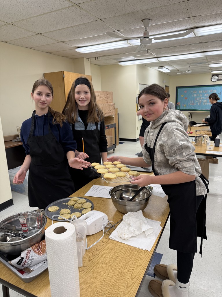 Three students wearing aprons stand at a table in a kitchen classroom holding a tray of freshly baked cookies, with mixing bowls and baking tools on the table.
